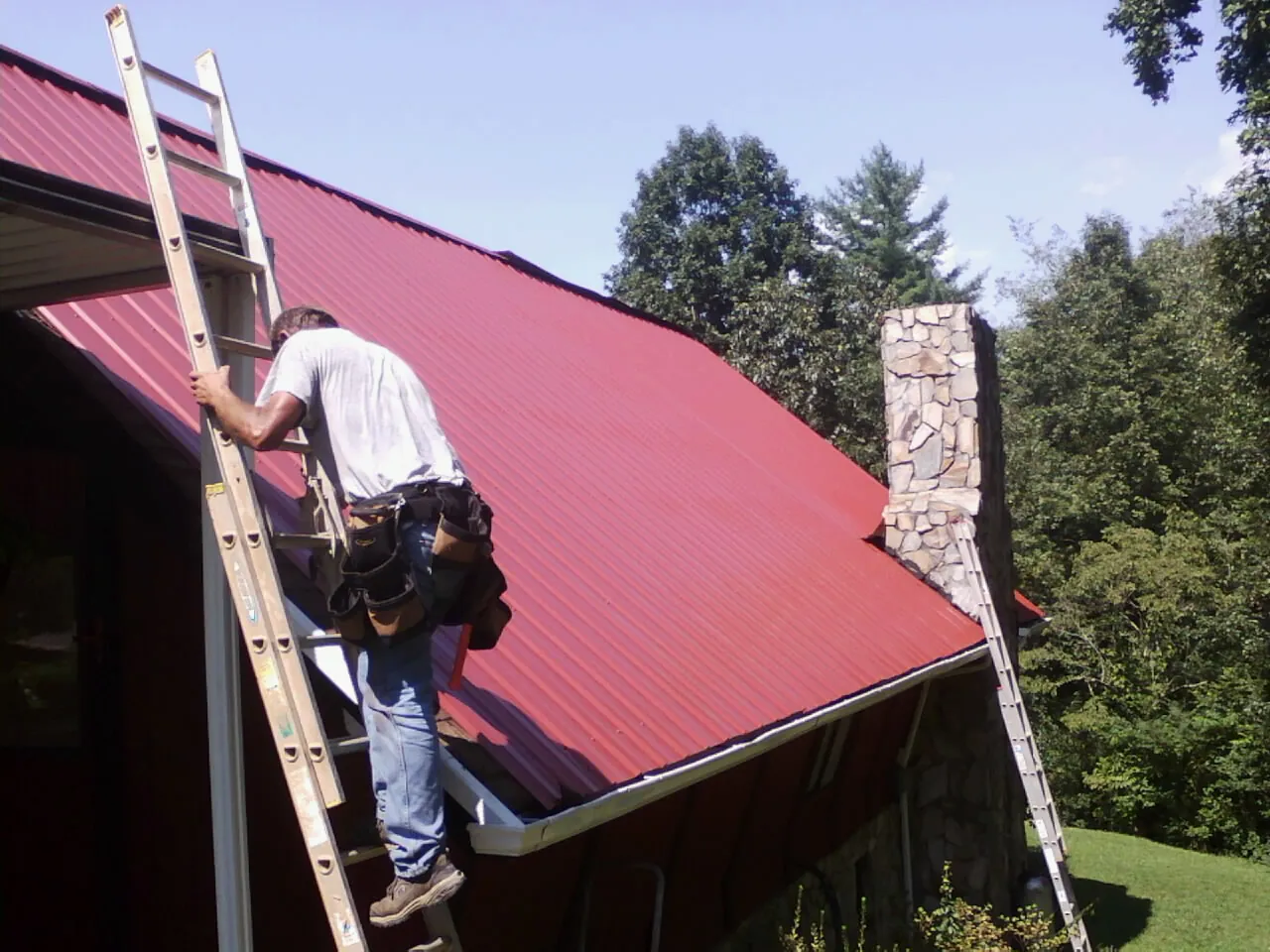 Roofer on a ladder beside a red metal roof and stone chimney — Soffit & Fascia Repair by Roofing Brothers in Bearcreek